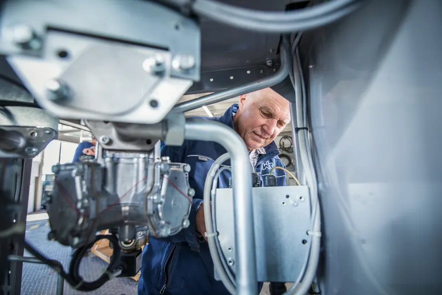 Im Vordergrund sind mechanische Komponenten und Rohrleitungen in einem technischen oder industriellen Umfeld zu sehen. Eine Person ist im Hintergrund teilweise von der Anlage verdeckt.