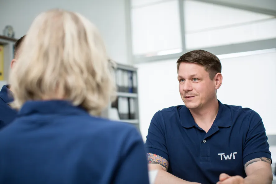 Zwei TWT-Mitarbeiter*innen in blauen Shirts mit dem TWT-Logo befinden sich in einem Gespräch in einem Besprechungsraum